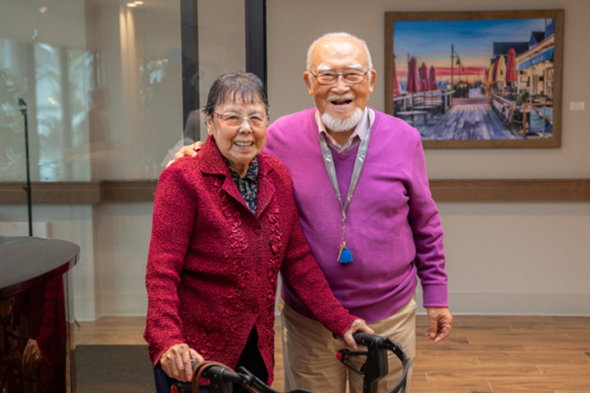 An older Asian couple with their arms around each other smiling at the camera