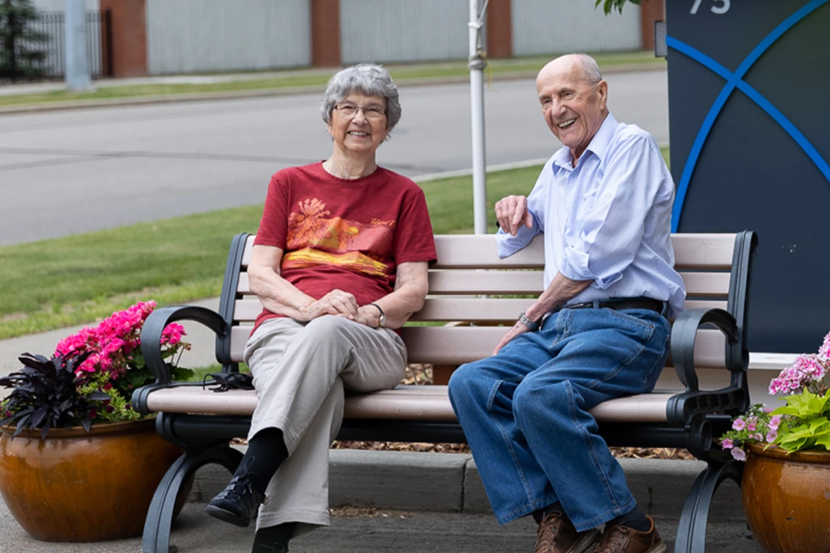 A senior couple sitting on a bench beside potted flowers outside a retirement home