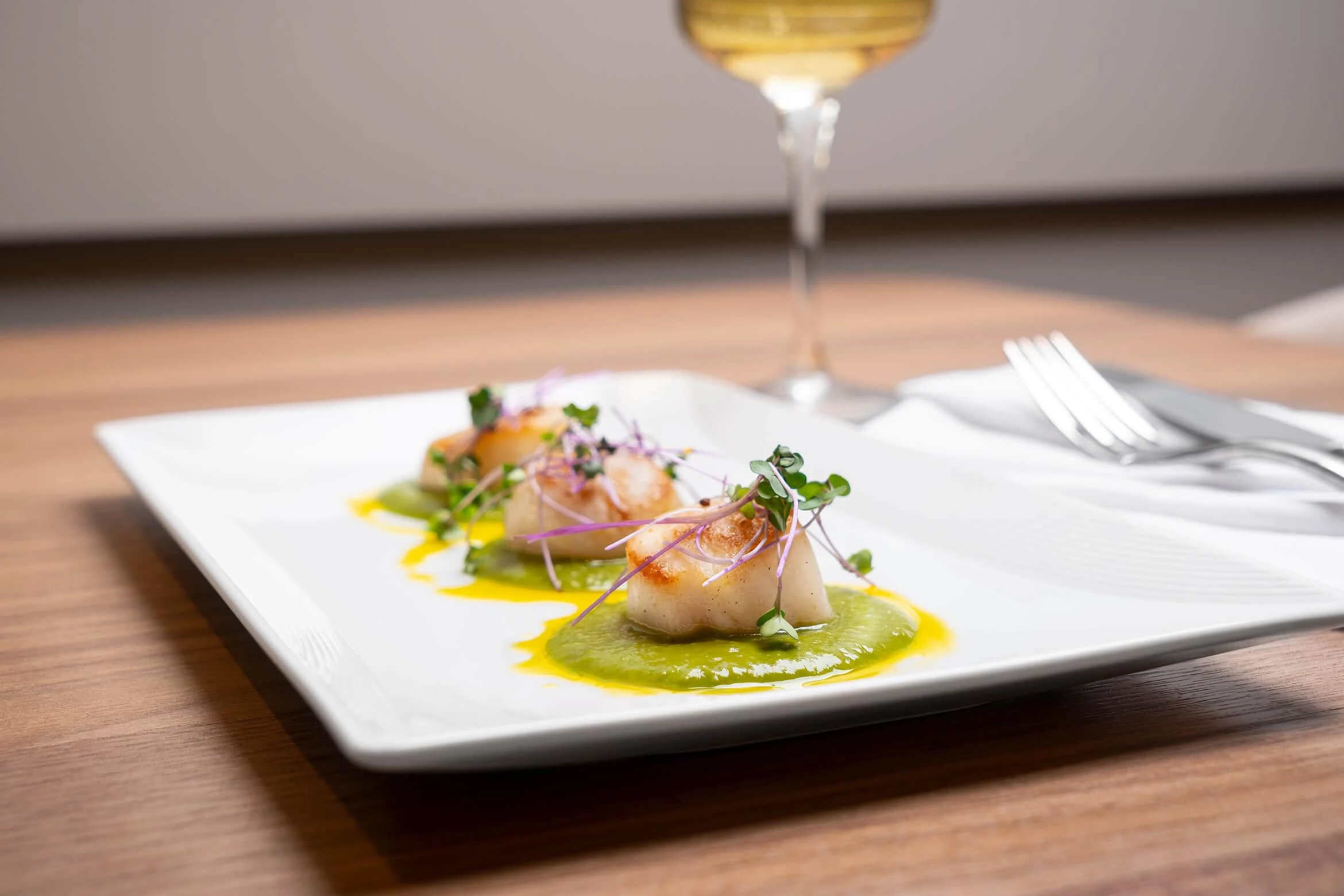 A plate of three scallops on top of green puree beside a glass of white wine.